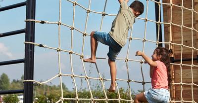 Banner mit zwei Kindern, die auf einem BERG PlayBase Klettergerüst mit Netz im Garten klettern