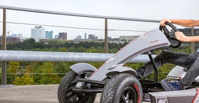 Boy driving a BERG Race XL pedal go-kart on an elevated bridge with a city view, banner