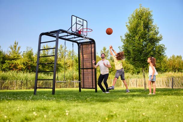 Conjunto de parque infantil BERG PlayBase com cesto de basquete e baloiços, onde um pai brinca ao ar livre com os filhos