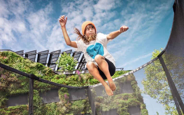 Enfant sautant sur un trampoline BERG avec filet de sécurité devant un mur végétalisé en ville