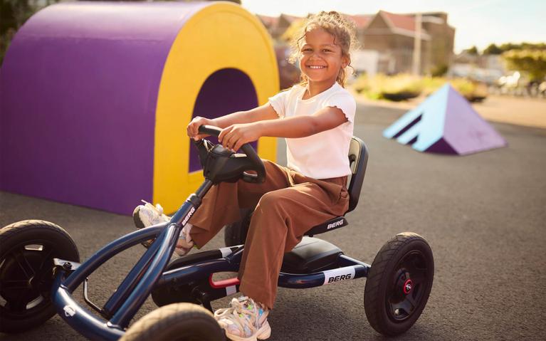 Smiling girl on a blue BERG Buddy pedal kart in a colorful play area
