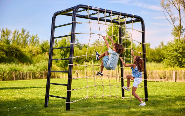 To barn klatrer på et BERG PlayBase lekestativ med nett på en solrik plen