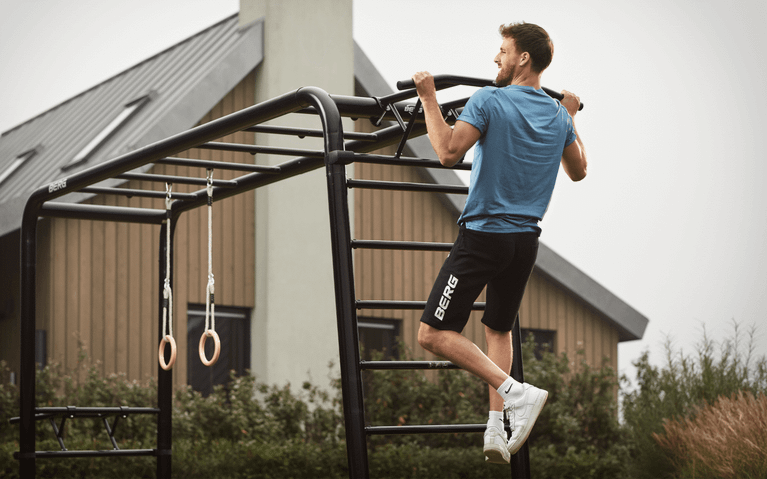 Mann macht Klimmzüge an der BERG PlayBase Klettergerüst mit Pull-up Bar im modernen Garten