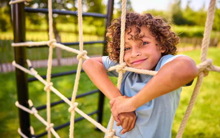 Gutt lener seg smilende mot nettet på et BERG PlayBase lekestativ på en solrik plen