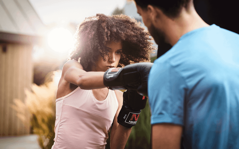 Frau trainiert mit Boxhandschuhen im Garten, unterstützt von einem Trainer am BERG PlayBase Klettergerüst