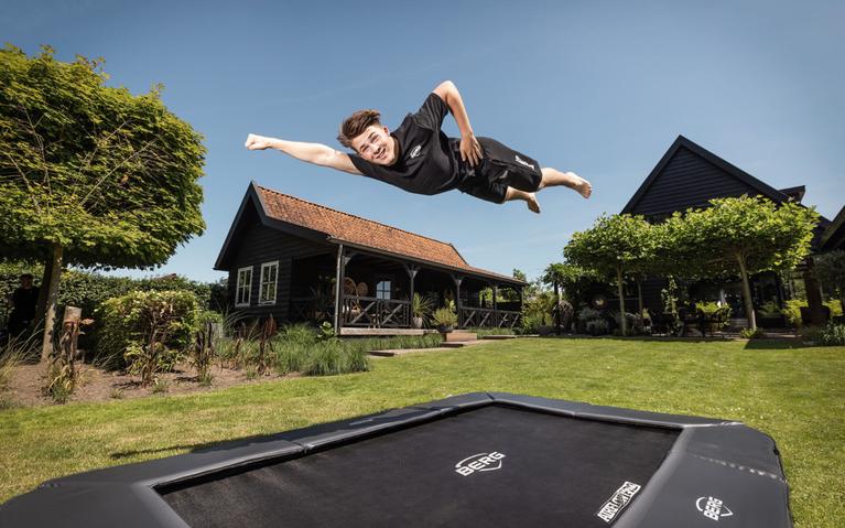 Bannière ludique avec garçon faisant un saut de super-héros sur trampoline BERG enterré dans un jardin