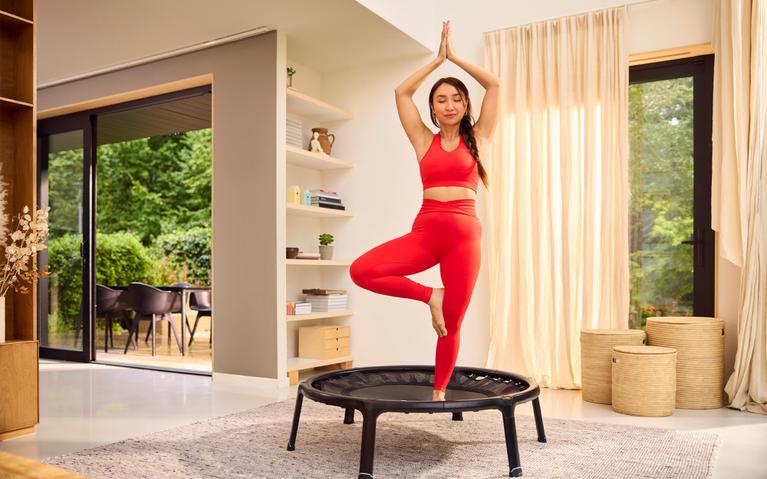 Frau steht in Yoga-Pose auf einem schwarzen BERG Fitness-Trampolin – Gleichgewichtsübung für zu Hause.