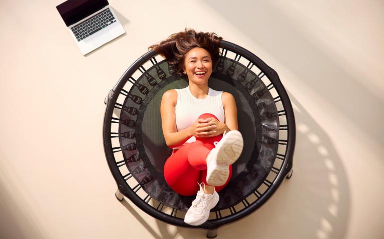 Frau liegt lachend auf einem schwarzen BERG Fitness-Trampolin nach dem Training – Entspannung zu Hause.