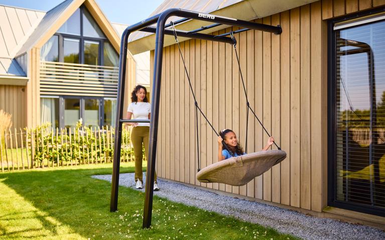 Mädchen entspannt in einer Nestschaukel am BERG PlayBase Klettergerüst neben einem Holzhaus