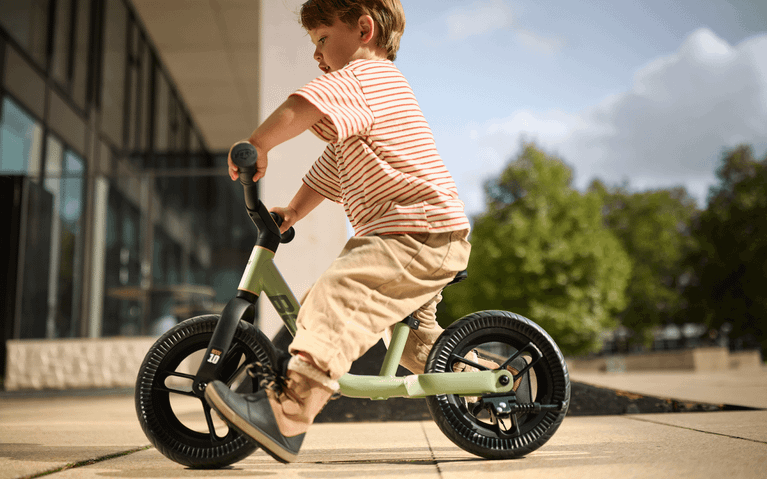 Kleinkind fährt auf einem grünen BERG Laufrad über einen gepflasterten Platz bei einem modernen Gebäude