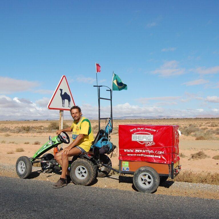 Pedal Joe with his BERG pedal kart and trailer on a desert road near a camel warning sign