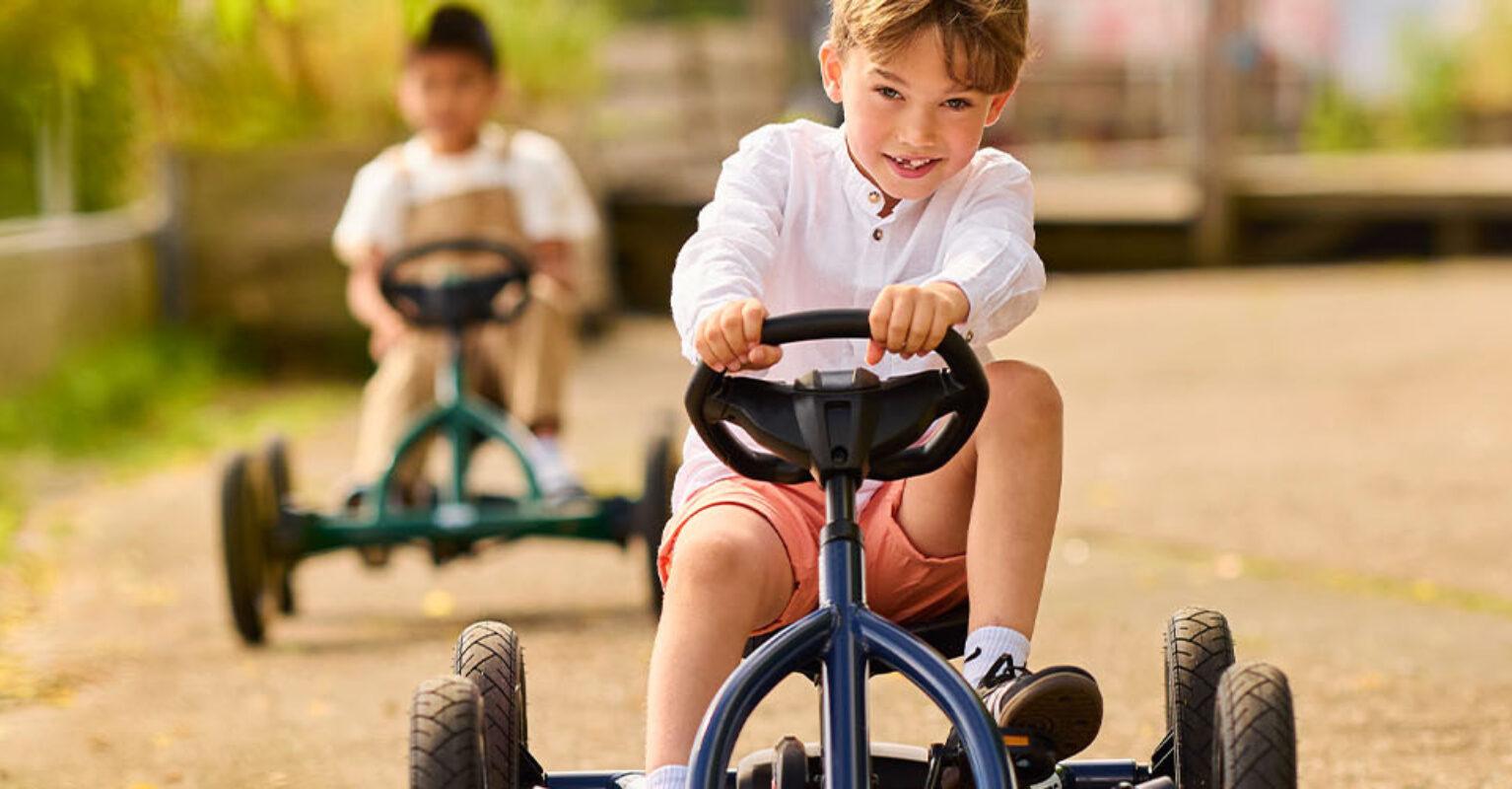 Bannière avec un garçon sur un kart à pédales BERG Buddy bleu sur un chemin ensoleillé suivi d’un ami