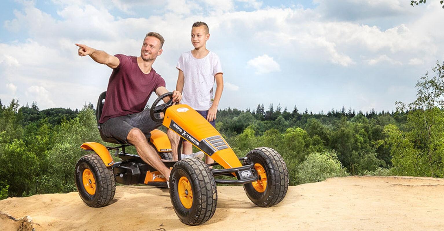 Banner mit zwei Jugendlichen auf einem orangefarbenen BERG Gokart auf einem Hügel mit Waldblick