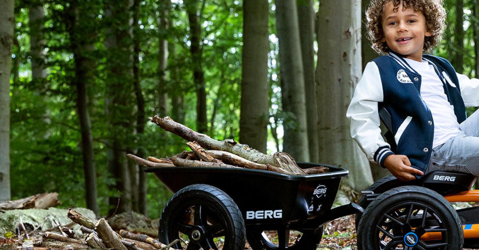 Banner mit Kind auf einem orangefarbenen BERG Buddy Gokart mit Anhänger voller Äste im Wald