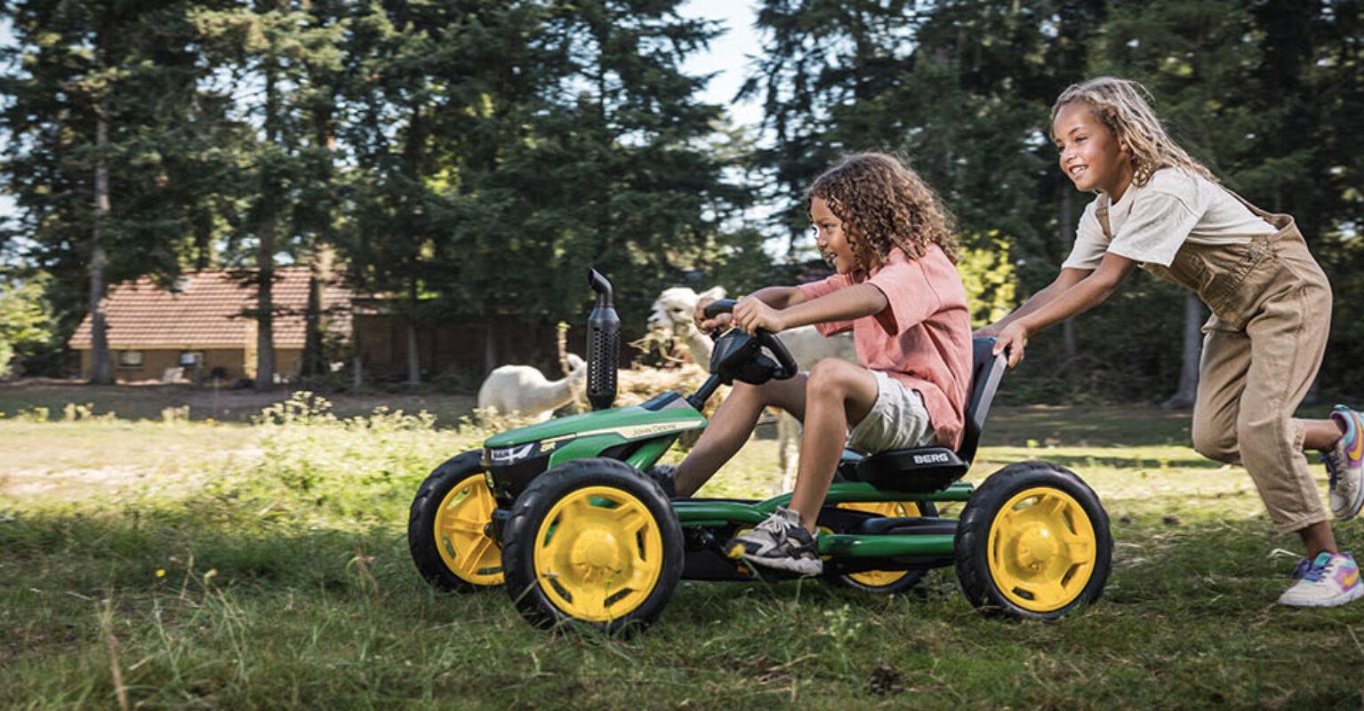 Meisje duwt een BERG John Deere skelter met een ander meisje erin over een grasveld